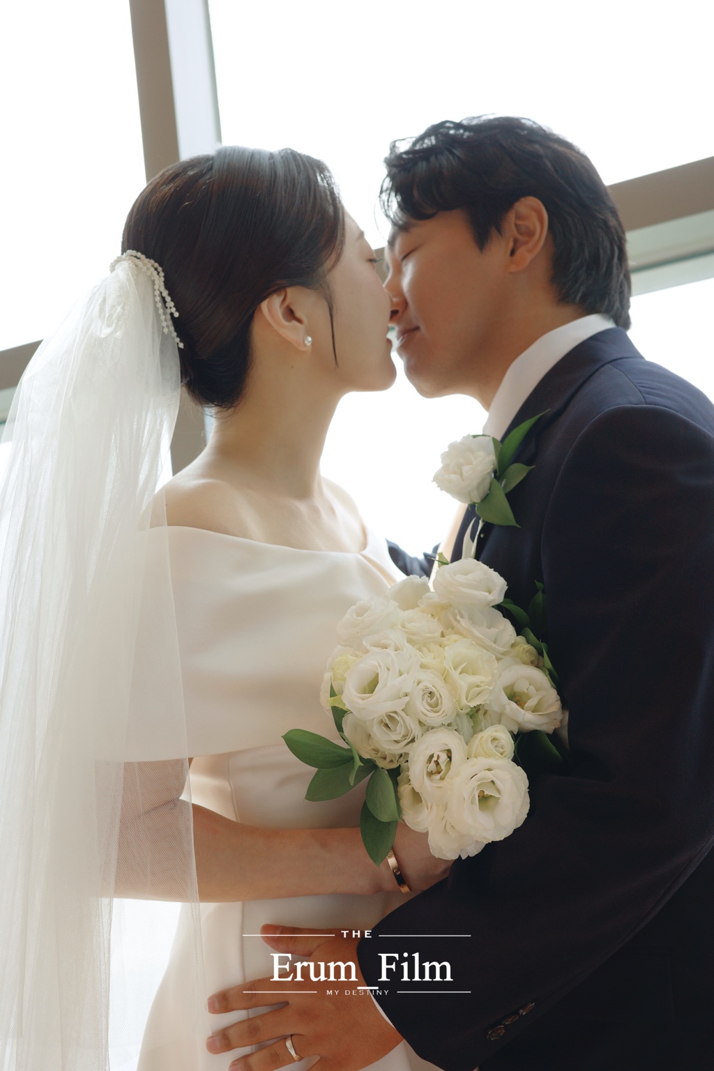 창가에서 꽃다발을 들고 입맞춤하는 엘리아 스몰웨딩 커플